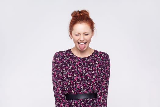 Portrait of naughty playful ginger woman with bun hairstyle wearing dress showing tongue at camera with closed eyes, having fun, joke. Indoor studio shot isolated on gray background.