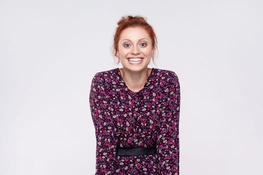 Portrait of extremely happy optimistic redhead woman wearing dress looking at camera with toothy smile, rejoicing, being in good mood. Indoor studio shot isolated on gray background.