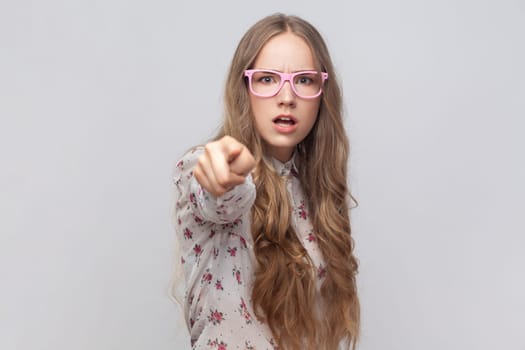 Portrait of angry serious young adult woman in glasses with wavy blond hair looking and pointing finger at camera, suspecting you. Indoor studio shot isolated on gray background.