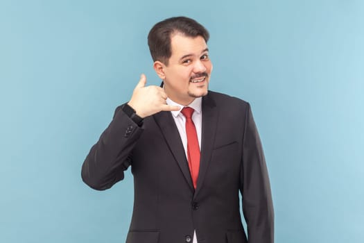 Please, call me back. Portrait of optimistic cute smiling man with mustache standing and showing call me gesture, wearing black suit with red tie. Indoor studio shot isolated on light blue background.