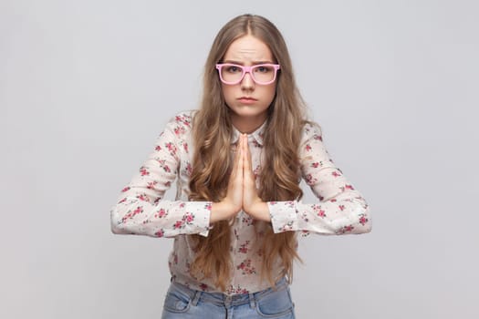 Please, need your help. Portrait of woman in glasses with long blond hair holding hands in praying gesture, pleading with desperate face. Indoor studio shot isolated on gray background.