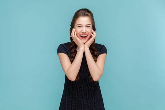 Portrait of funny joyful cheerful woman with braids standing with hands under chin and laughing, hearing joke, wearing black dress. woman Indoor studio shot isolated on blue background.