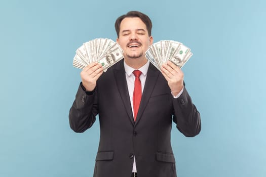 Portrait of greedy smiling man with mustache standing with closed eyes, holding dollar banknotes in hands, wearing black suit with red tie. Indoor studio shot isolated on light blue background.