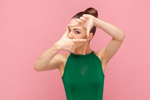 Portrait of concentrated woman photographer with bun hairstyle standing and looking at camera, making frame with fingers, wearing green dress. Indoor studio shot isolated on pink background.