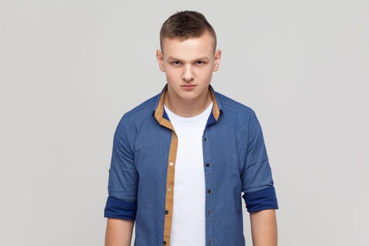 Portrait of enraged furious attractive teenager boy wearing blue shirt standing looking at camera, expressing wild anger and hate, feeling crazy. Indoor studio shot isolated on gray background.