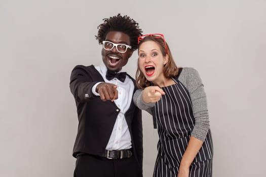 Portrait of excited happy positive woman and man in white glasses standing pointing at you, laughing out loud, shame on you. Indoor studio shot isolated on gray background.