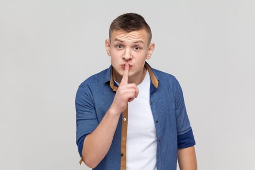 Please, be quiet. Teenager boy in blue shirt shushing with silence gesture, holding finger on lips and smiling to camera, asking to keep secret. Indoor studio shot isolated on gray background.