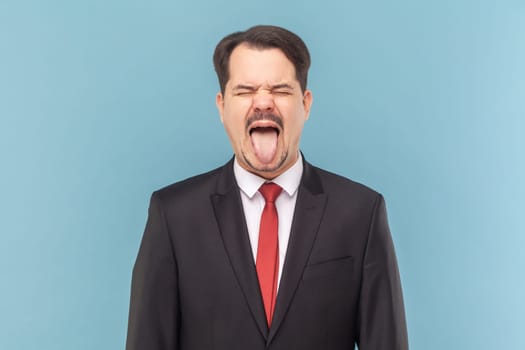 Portrait of crazy excited man with mustache standing sticking tongue out, demonstrates childish behavior, wearing black suit with red tie. Indoor studio shot isolated on light blue background.