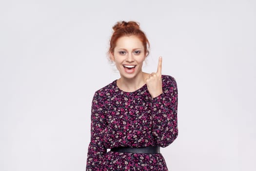 Portrait of astonished excited redhead woman wearing dress pointing finger up with genius idea, surprised by suddenly invented smart solution. Indoor studio shot isolated on gray background.