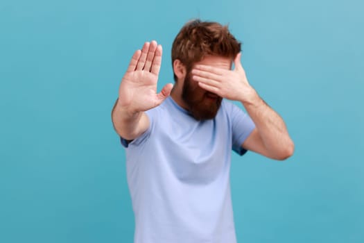 Portrait of bearded man standing, closed his eyes with hands and showing stop gesture, feeling stressed afraid, refusing to watch scary content. Indoor studio shot isolated on blue background.