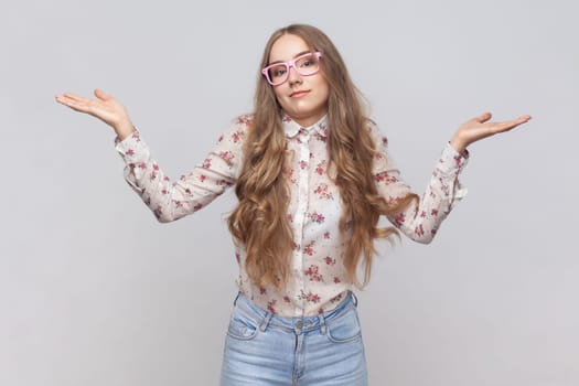 I don't know. Portrait of confused beautiful woman in glasses with wavy blond hair standing, raised arms, confused and thinking. Indoor studio shot isolated on gray background.