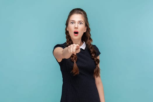 Portrait of shocked astonished woman with braids standing pointing to camera, sees something amazing, being surprised, wearing black dress. woman Indoor studio shot isolated on blue background.