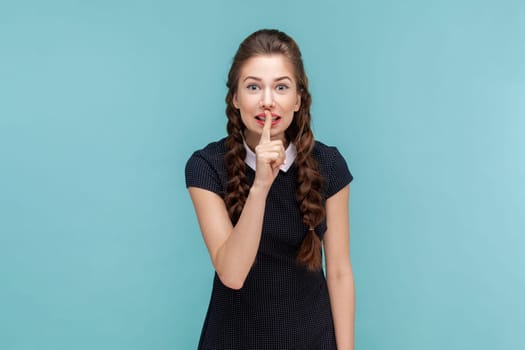 Portrait of smiling optimistic attractive woman with braids standing with finger near lips, asking to keep secret, wearing black dress. woman Indoor studio shot isolated on blue background.