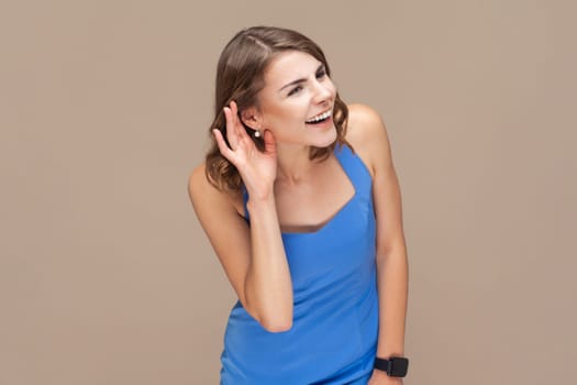 Portrait of attractive beautiful young adult woman with wavy hair standing palm near ear, listening private conversation, wearing blue dress. Indoor studio shot isolated on light brown background.