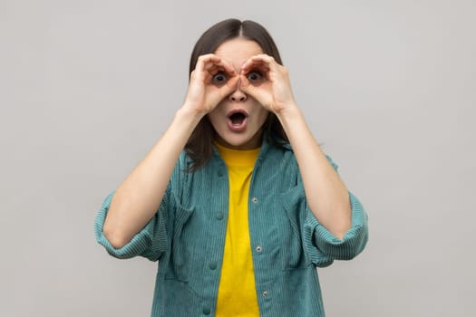 Shocked attractive young woman standing with hands on eyes binoculars gesture and looking at camera with surprised face, wearing casual style jacket. Indoor studio shot isolated on gray background.