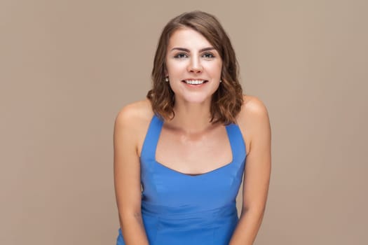 Portrait of smiling positive happy woman with wavy hair looking at camera with toothy smile, being in good mood, being glad, wearing blue dress. Indoor studio shot isolated on light brown background.