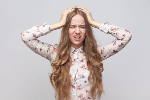 Portrait of unhealthy sick young adult woman with wavy blond hair holding hands on head, feeling terrible headache, risk of migraine. Indoor studio shot isolated on gray background.