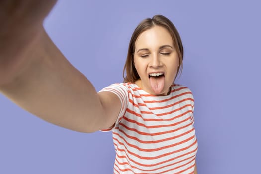 Portrait of funny childish woman in striped T-shirt taking selfie picture, point of view, showing tongue out to camera, making front selfportrait. Indoor studio shot isolated on purple background.