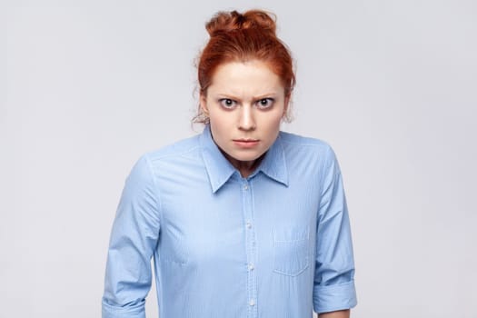 Portrait of angry aggressive ginger woman wearing blue shirt looking at camera with anger and hate, being in bad nervous mood. Indoor studio shot isolated on gray background.