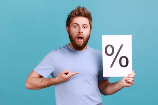 Portrait of bearded man holding paper with percent sign inscription, looking at camera with open mouth and shocked expression. Indoor studio shot isolated on blue background.