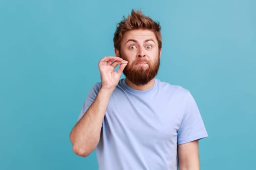 I don't tell anyone. Portrait of handsome young adult bearded man zipping mouth, keeping secret information, looking at camera. Indoor studio shot isolated on blue background.