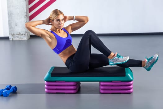 Side view portrait of sporty beautiful woman with blonde hair wearing tight sportswear sitting on step platform and doing press aerobic, having work out in fitness club. Indoor shot.