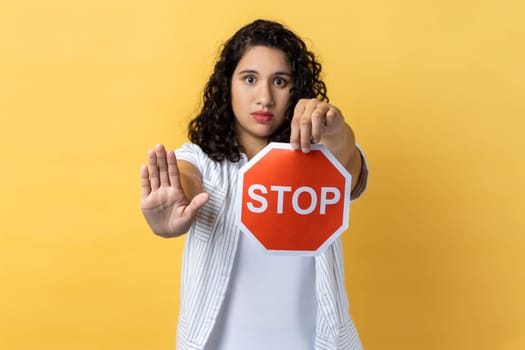 Portrait of strict woman with dark wavy hair holding red stop sign looking at camera with ban palm gesture, has strict expression, prohibition. Indoor studio shot isolated on yellow background.