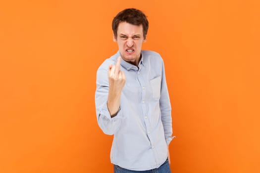 Portrait of crazy man shows fuck you sign looking with poker face being vulgar and has quarrel with someone, wearing light blue shirt. Indoor studio shot isolated on orange background.