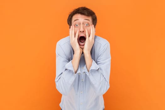 Portrait of shocked scared man looking at camera with open mouth, keeps hands on cheeks, being frighten, wearing light blue shirt. Indoor studio shot isolated on orange background.