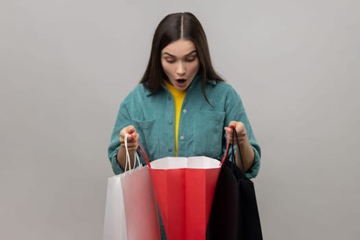 Excited amazed woman with dark hair looking inside shopping bags, having shocked expression, being surprised, wearing casual style jacket. Indoor studio shot isolated on gray background.