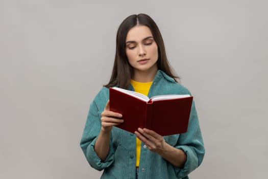 Portrait of serious concentrated woman with dark hair reading book with interesting plot, studying, wearing casual style jacket. Indoor studio shot isolated on gray background.