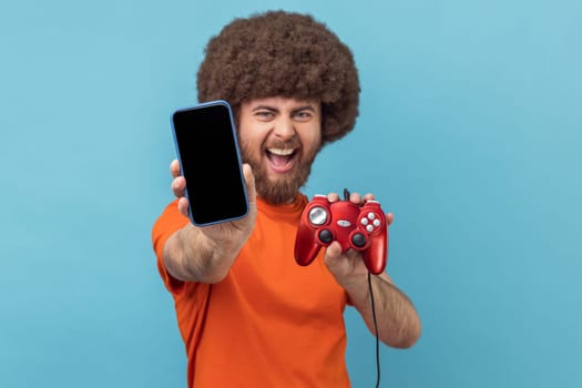 Portrait of excited man with Afro hairstyle wearing orange T-shirt holding out showing joypad and showing mobile phone with empty display. Indoor studio shot isolated on blue background.