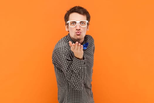 Portrait of funny positive man nerd sending air kissing, falling in love, flirting, wearing shirt with blue bow tie and white glasses. Indoor studio shot isolated on orange background.