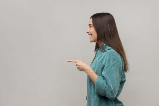 Side view of happy beautiful young adult woman standing, pointing and showing copyspace with happy face and smile, wearing casual style jacket. Indoor studio shot isolated on gray background.