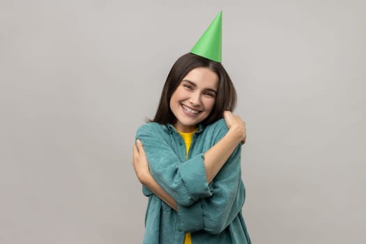 I love myself. Woman with dark hair and funny party cone on head, congratulating and embracing herself on birthday, wearing casual style jacket. Indoor studio shot isolated on gray background.