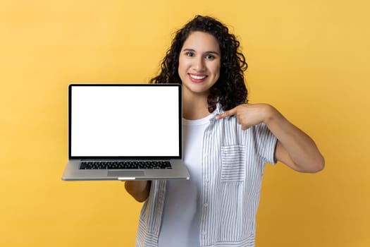 Portrait of delighted optimistic woman with dark wavy hair holding and pointing laptop with empty display, copy space for advertisement. Indoor studio shot isolated on yellow background.