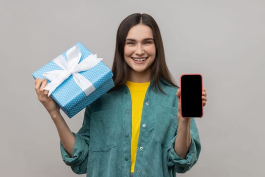 Present, bonus for mobile user. Woman holding gift box and cell phone with empty display for online shopping advertising, wearing casual style jacket. Indoor studio shot isolated on gray background.