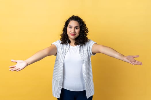 Please, take for free. Portrait of generous friendly woman with dark wavy hair welcoming with wide open arms and smiling kindly, happy to embrace you. Indoor studio shot isolated on yellow background.