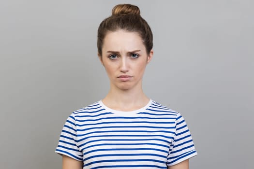 Portrait of sad upset young adult woman wearing striped T-shirt expressing sadness, hearing bad news, being in bad mood. Indoor studio shot isolated on gray background.