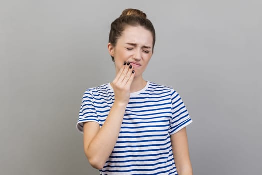 Tooth ache. Portrait of woman wearing striped T-shirt standing and holding her painful tooth, frowning face from terrible pain. Indoor studio shot isolated on gray background.