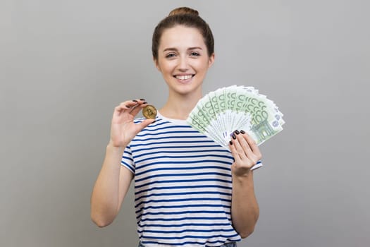 Portrait of delighted positive woman wearing striped T-shirt holding golden coin of crypto currency and big fan of euro banknotes. Indoor studio shot isolated on gray background.