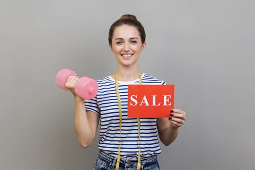 Portrait of smiling happy woman wearing striped T-shirt holding pink dumbbell and card with sale inscriptions, discounts for gym membership. Indoor studio shot isolated on gray background.