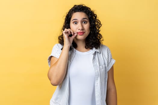 Portrait of attractive woman with dark wavy hair and mystery look making zip gesture to close mouth, keeping secret, zipping lips. Indoor studio shot isolated on yellow background.