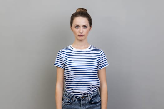 Portrait of beautiful serious bossy woman with bun hairstyle wearing striped T-shirt standing looking at camera with strict facial expression. Indoor studio shot isolated on gray background.