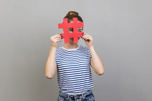 Portrait of unknown woman wearing striped T-shirt looking through red hashtag sign, looking for proper posts in social media, spying. Indoor studio shot isolated on gray background.