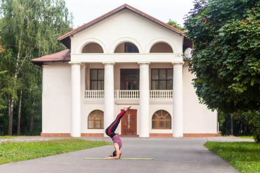 Full length side view of young strong sporty woman doing yoga asana supported headstand on the street, Salamba Sirsasana, complex balanced posture for a healthy body and spirit.