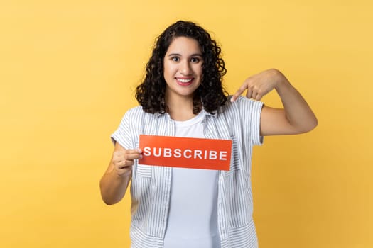 Portrait of cheerful young adult woman with dark wavy hair holding and pointing finger at red card with subscribe inscription. Indoor studio shot isolated on yellow background.