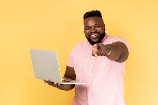 Portrait of optimistic bearded man wearing pink shirt holding laptop, pointing finger to camera and smiling, choosing you, freelance job advertisement. Indoor studio shot isolated on yellow background