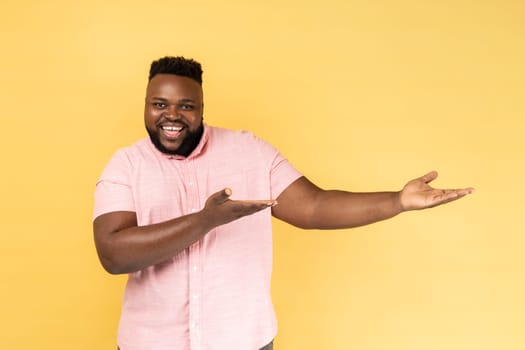 Please, take for free. Portrait of man wearing pink shirt welcoming with wide open arms and smiling kindly, looking at camera with toothy smile. Indoor studio shot isolated on yellow background.