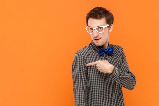 Portrait of man nerd pointing away on copy space, discusses amazing promo, gives way or direction, wearing shirt with blue bow tie and white glasses. Indoor studio shot isolated on orange background.